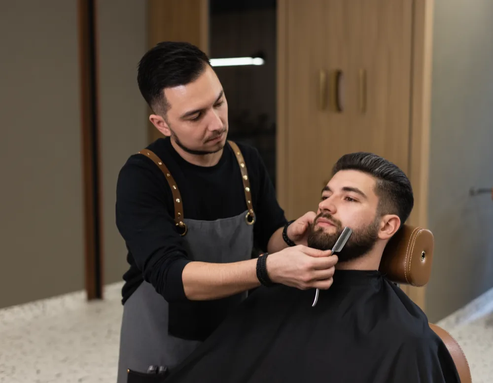 A barber shaping a short full beard along the cheek line with a straight razor