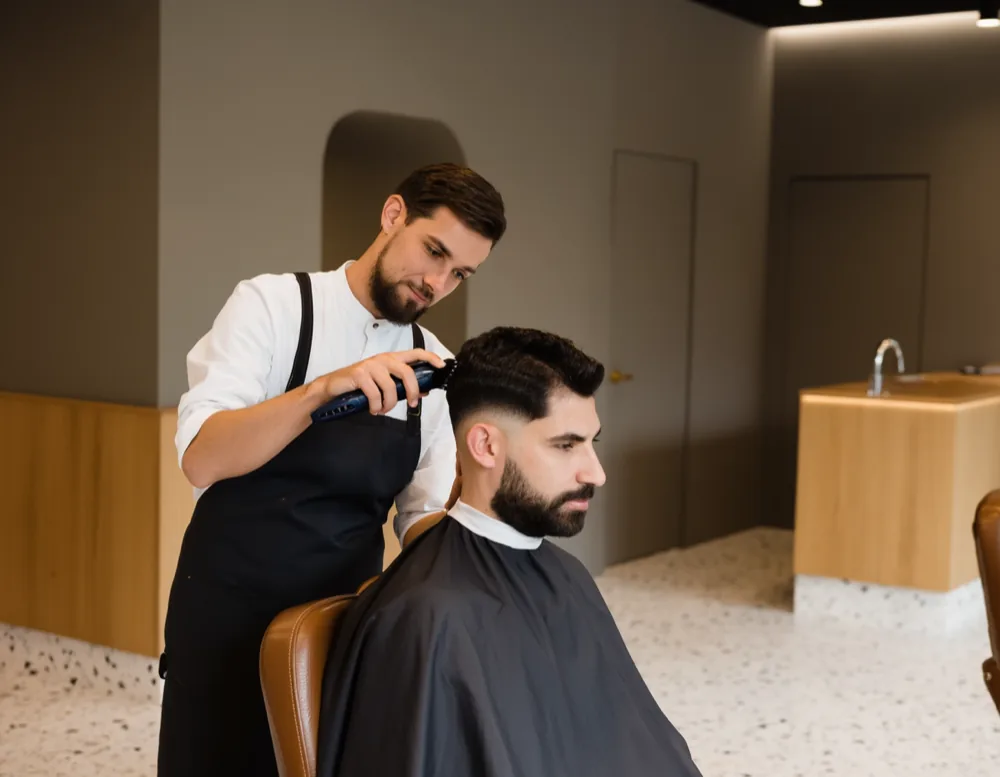 A barber in a charcoal apron cutting the side of a seated client’s hair with clippers